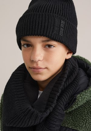 Jonge persoon met een zwarte geribbelde gebreide beanie en bijpassende sjaal met de tekst "Blue Ridge", gekleed in een groene en zwarte fleecejas.