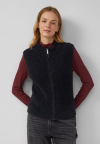 Schwarze, gerippte Strickweste mit hohem Kragen und vorderem Reißverschluss, getragen über einem kastanienbraunen Langarmshirt. Weicher Stoff und taillierte Passform.