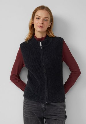 Schwarze, gerippte Strickweste mit hohem Kragen und vorderem Reißverschluss, getragen über einem kastanienbraunen Langarmshirt. Weicher Stoff und taillierte Passform.