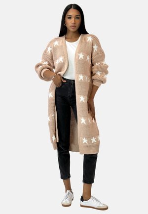 Cardigan lungo in maglia di un leggero rosa con motivi di stelle bianche, con apertura frontale e maniche oversize, abbinato a una maglietta bianca e jeans scuri.