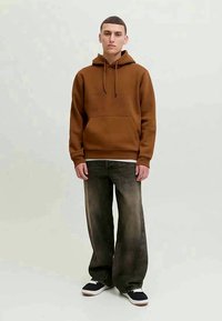 Young man standing, wearing brown hoodie, loose faded black jeans, and black sneakers with white soles.