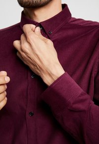 Bordeauxrotes Kordhemd mit Button-Down-Kragen, schwarzen Knöpfen und gerippter Textur, Hände, die den Kragen anpassen.