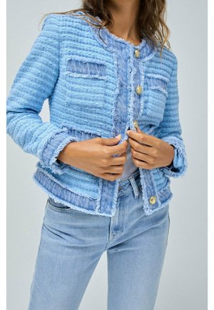 Veste bleu clair texturée avec des bords effilochés, deux poches avant et des boutons dorés ; portée sur une chemise claire et associée à un jean bleu.