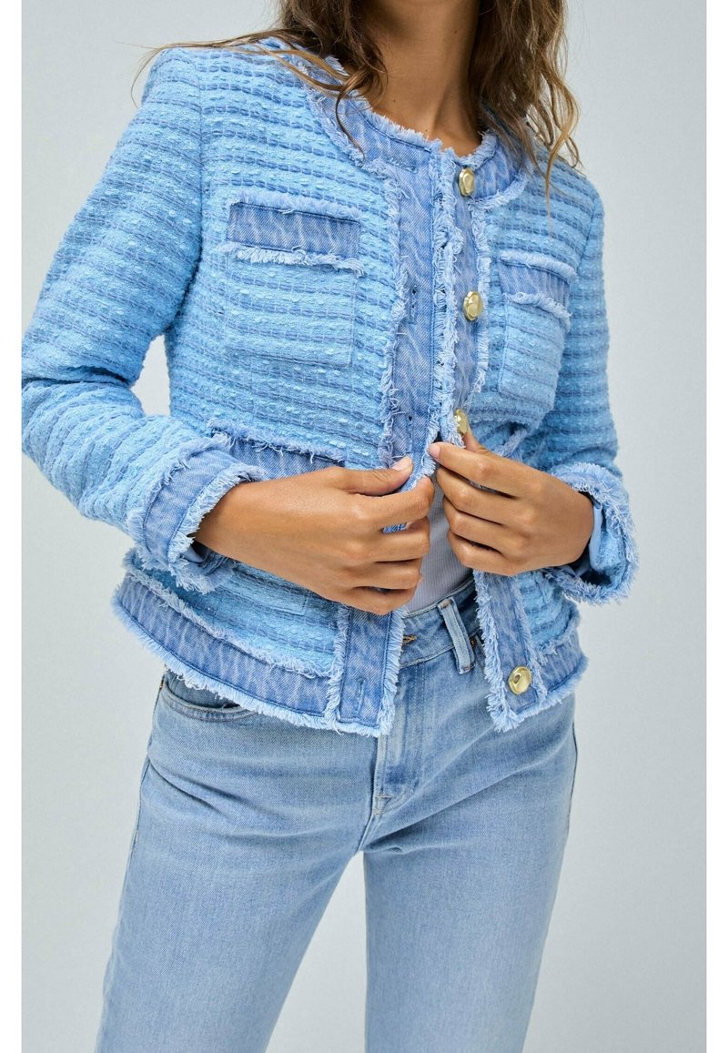 Ljusblå, strukturerad jacka med fransade kanter, två framfickor och guldknappar; bärs över en ljus skjorta och kombineras med blå jeans.