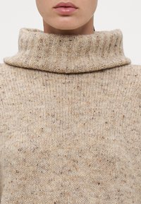 Beige gebreide trui met een hoge, geribbelde col. De textuur van de stof heeft verspreide donkere vlekken door het materiaal.