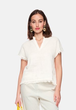 Woman with shoulder-length brown hair wears white fringed short-sleeve top, white pants, large gold earrings, holding a yellow beaded handbag.