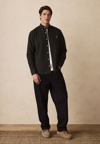 Čierna košeľa na gombíky cez biele tričko, v kombinácii s tmavými džínsami a béžovými semišovými slip-on topánkami, na neutrálnom pozadí.