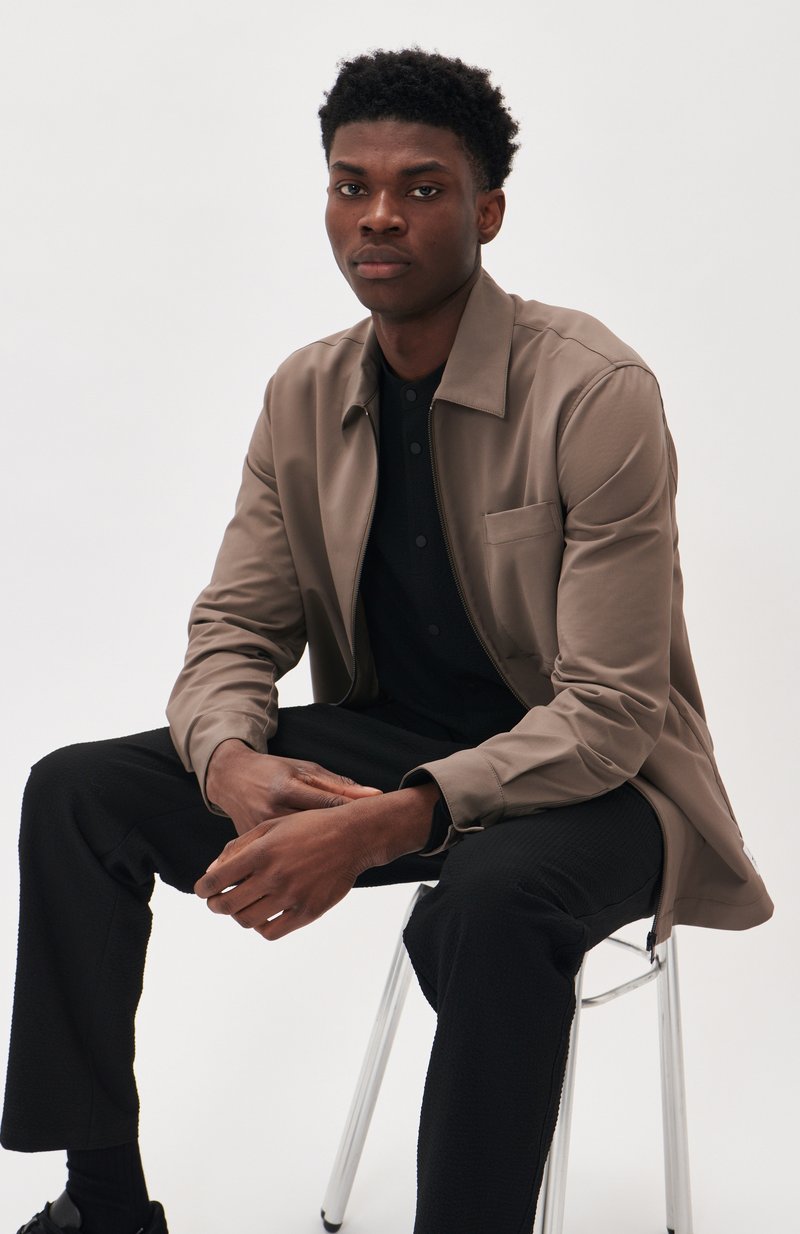Veste zippée beige clair avec un col, portée sur une chemise noire à boutons et un pantalon noir texturé, assis sur une chaise argentée.