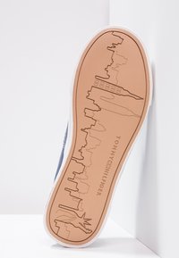 Sula på skon med en tan gummi-bas präglad med en stadsprofil och Frihetsgudinnan. Sidan har en vit kant med mörkblått ovandelsmaterial.