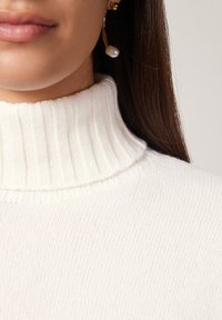 Weißer gerippter Rollkragenpullover mit einer weichen Textur. Goldene Creolen mit einem Perlenakzent. Nahaufnahme des Ausschnitts und des unteren Gesichts.