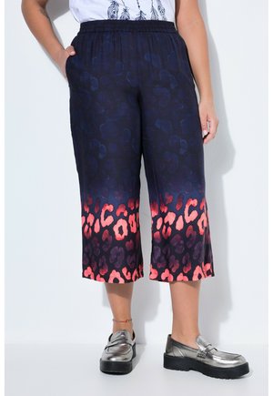 Vrouw draagt cropped broek met donkerblauwe en rode luipaardprint, een witte top en zilveren platform loafers, staand tegen een witte achtergrond.