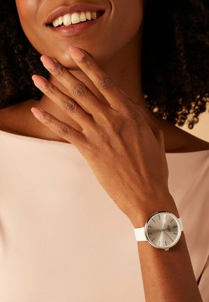 Nahaufnahme einer lächelnden Person mit brauner Haut, die eine silberne Uhr mit weißem Armband und passenden mauvefarbenen Nagellack trägt.