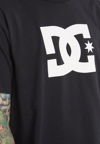Černé tričko s výrazným bílým logem DC a hvězdným akcentem. Tkanina vypadá měkce a má klasický kulatý výstřih.