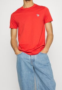 Camiseta de algodón roja con cuello redondo y mangas cortas; presenta un pequeño gráfico de cebra en el lado izquierdo del pecho; combinada con jeans de color azul claro.