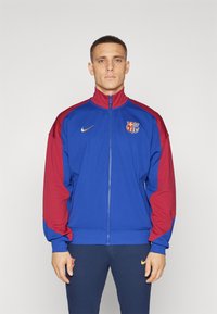 Mann med blå og rød FC Barcelona-jakke med glidelås, kombinert med marineblå bukser med gull Nike og klubblogoer.