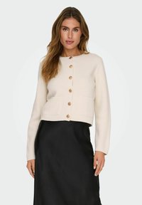 Cardigan a maglia color crema con scollo rotondo, caratterizzato da sei bottoni in legno e due tasche anteriori. Indossato sopra una gonna nera.