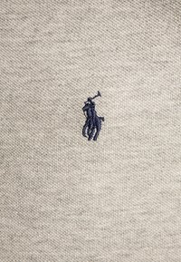 Světle šedá pletenina svetr s texturovaným vzorem, na levé části hrudi je malá tmavě modrá výšivka loga koně a jezdce.