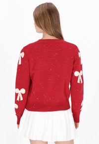 Pullover rosso in maglia con motivi a fiocco bianchi sulle maniche, dettagli lucidi sottili e orlo a coste. Maniche lunghe e scollo a giro.