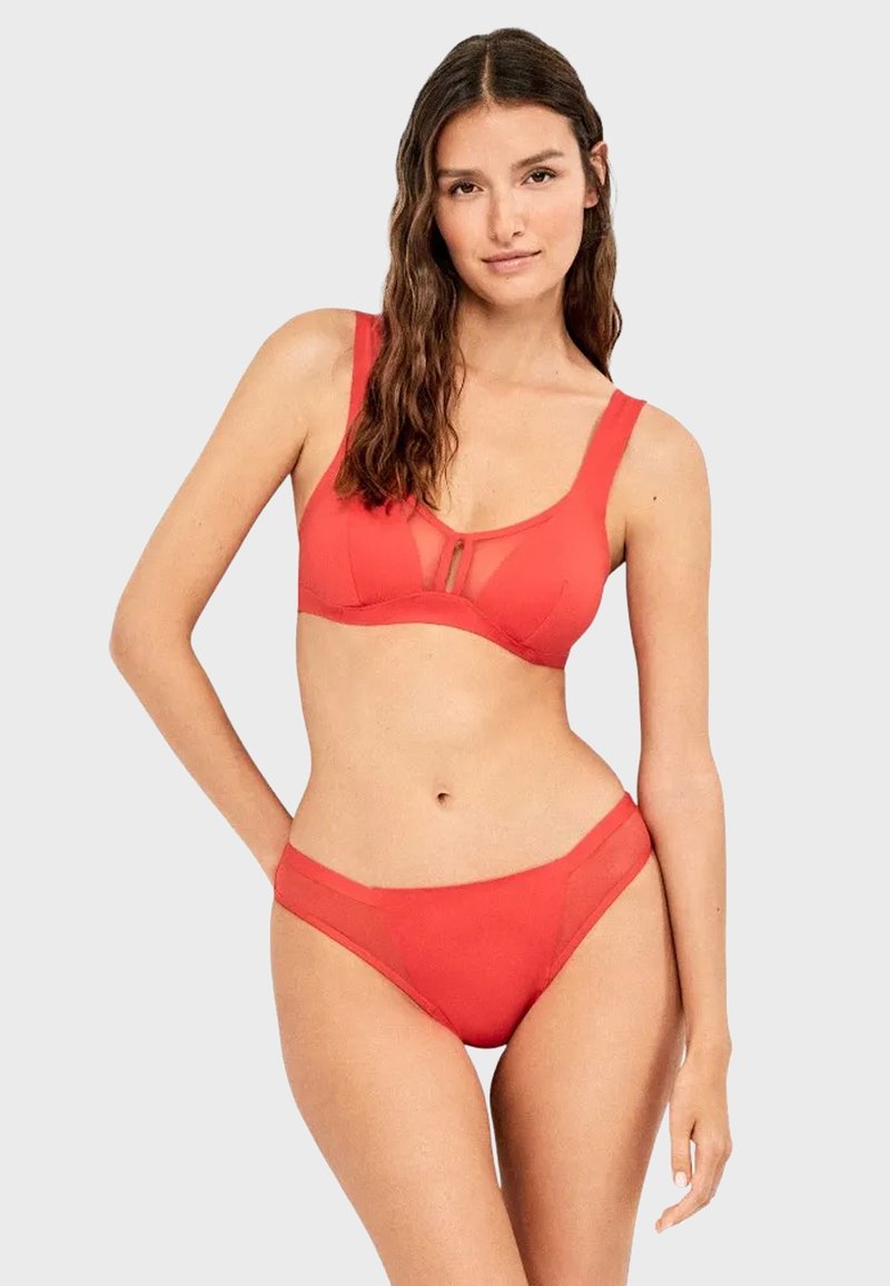 Femme aux longs cheveux bruns portant un ensemble de lingerie rouge deux pièces avec des détails en résille, debout devant un fond clair uni.