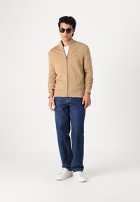 Sweat à zip beige avec des poignets côtelés, porté sur un t-shirt blanc, associé à un jean droit bleu marine et des baskets blanches. Lunettes de soleil noires.