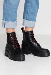 Botas de tobillo de cuero negro con cordones rojos y negros, suelas gruesas, cremallera lateral y detalles en capas. Llevadas con unos vaqueros claros con vuelta.