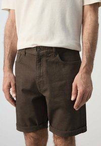 Hombre que lleva pantalones cortos de color marrón oscuro con dobladillo enrollado y una camisa de manga corta beige claro, con las manos relajadas a los lados.