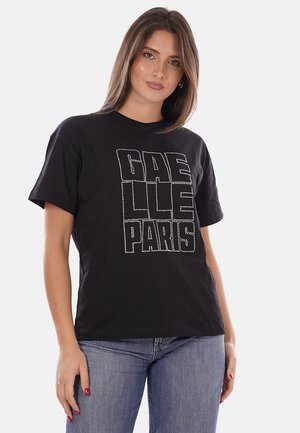 Femme portant un t-shirt noir avec "GAELLE PARIS" en lettres ponctuées, jean bleu, debout devant un fond gris clair uni.