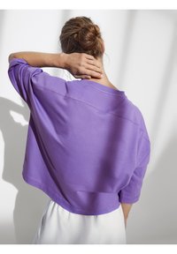 Felpa corta viola con vestibilità ampia e spalle scese. Texture liscia con dettagli di cuciture visibili. Indossata con pantaloni bianchi.