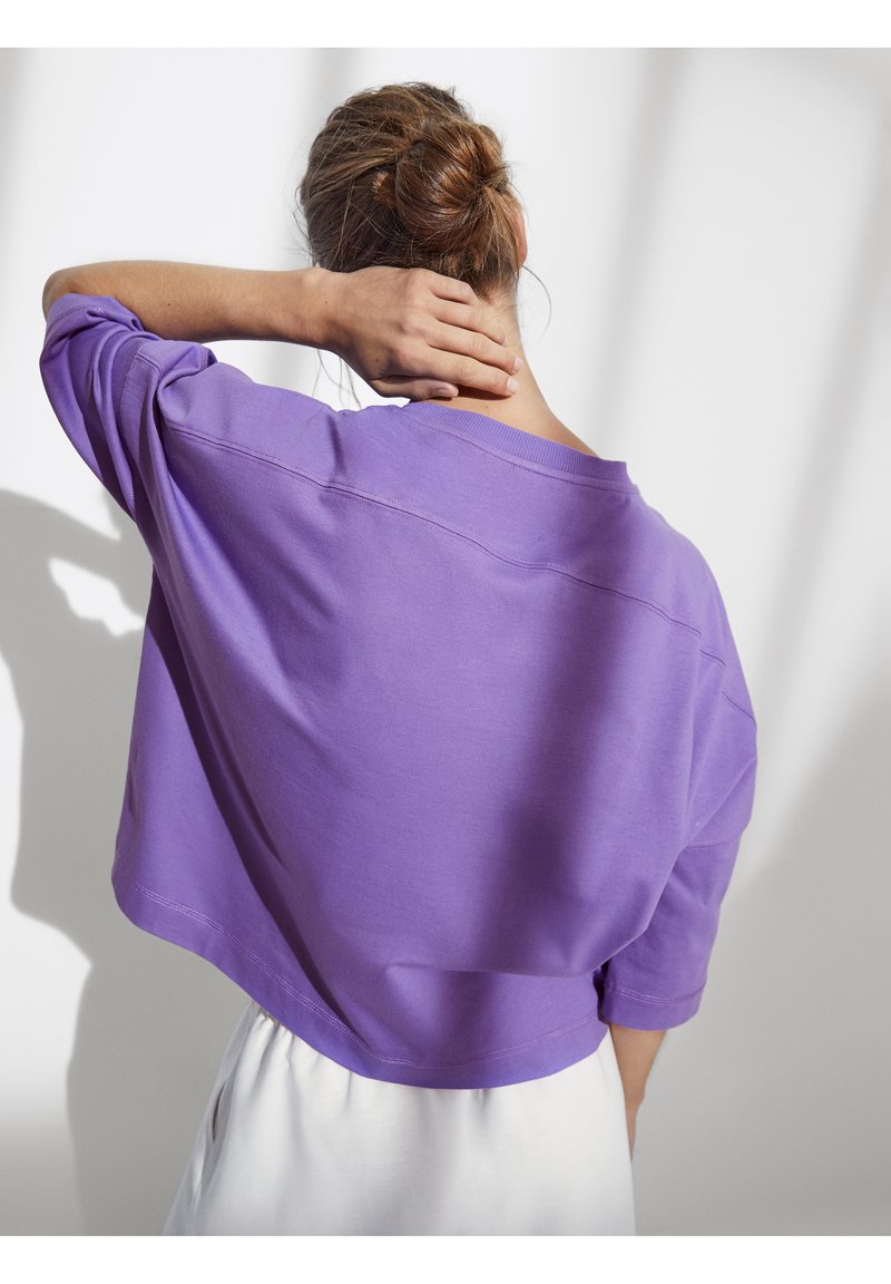 Felpa corta viola con vestibilità ampia e spalle scese. Texture liscia con dettagli di cuciture visibili. Indossata con pantaloni bianchi.