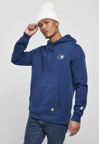 Felpa blu navy con tasca frontale, cappuccio regolabile con cordino e logo sul petto. Presenta polsini e orlo a coste. Indossata con jeans di denim.