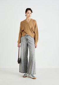 Kvinne iført en beige omslagsgenser, vidutsvingte falmede grå jeans, sølvfargede sneakers, og holder en liten brun håndveske mot en enkel hvit bakgrunn.