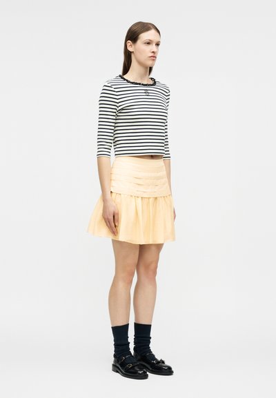Γυναίκα που στέκεται φορώντας ένα κοντό τοπ με μαύρες και λευκές ρίγες, ανοιχτό κίτρινο πλισέ φούστα, μαύρες κάλτσες και μαύρα μοκασίνια σε λευκό φόντο.
