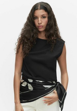 Vrouw met lang krullend haar, gekleed in een mouwloos zwart topje en een zwart-wit sjaal met patroon om haar taille gestrikt, witte broek zichtbaar.