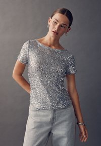 Top a maniche corte con paillettes argento e scollo ampio, abbinato a jeans grigio chiaro a vita alta. Leggero splendore e superficie strutturata.