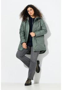 Grön vattentät jacka med huva, dragkedjefickor och mjuk insida, ihop med grå jeans och kraftiga olivgröna stövlar.