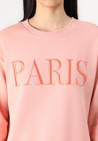 Rózsaszín pulóver hímzett "PARIS" felirattal, ugyanabban az árnyalatban. A szövet puha, bordázott környakú, laza fazonnal.