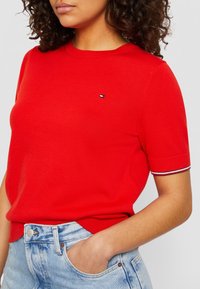 Czerwona dziana koszulka z krótkim rękawem, z małym logo na piersi i paskowanymi akcentami na mankietach rękawów. Noszona z niebieskimi szortami dżinsowymi.