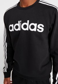 Sweat-shirt noir en coton avec un grand logo "adidas" blanc sur le devant. Des bandes blanches descendent le long des manches. Design simple.
