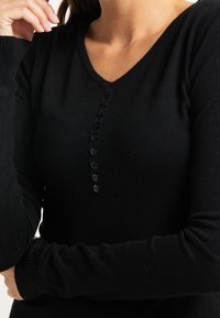 Maglione nero lavorato a maglia con scollo a V e maniche lunghe. Presenta una fila di bottoni neri lungo la parte anteriore per la chiusura. Texture morbida.