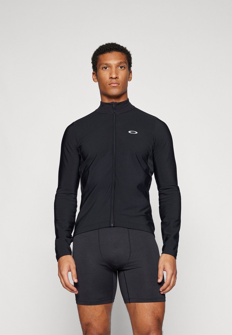 Black long-sleeve cycling jacket with a high collar and front zipper. Features a small logo on the chest. Worn with black cycling shorts.