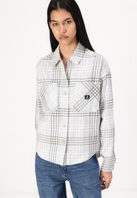 Camisa de botão xadrez em cinza claro e branco, com dois bolsos frontais e um pequeno emblema. Tecido suave com um corte descontraído.