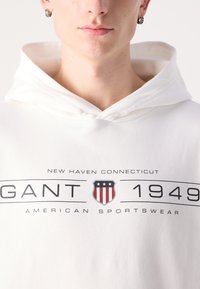 Felpa bianca in cotone con grafica "GANT 1949" e un emblema a scudo in rosso, blu e bianco, che mette in evidenza elementi di design sportivo.