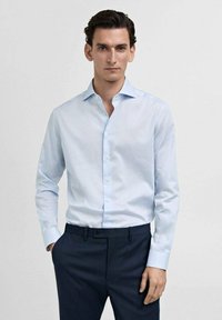 Cămașă de dress cu dungi albastre deschise, cu guler, mâneci lungi și închidere cu butoane, asortată cu pantaloni închiși la culoare. Textură de țesătură netedă.