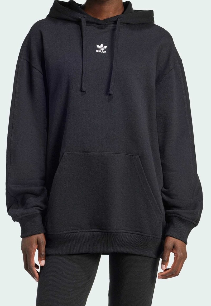 Svart hoodie med en stor passform, gjord av mjukt tyg. Har en framficka, dragsnören och en vit Adidas-logotyp på bröstet.