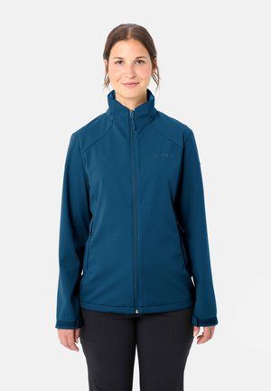 Frau trägt eine blaue Jacke mit Reißverschluss und hohem Kragen sowie schwarze Hose, steht vor einem schlichten hellen Hintergrund.