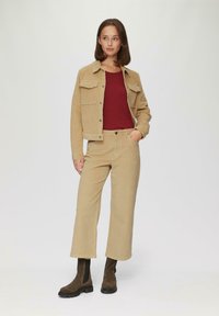 Beige corduroy jasje met twee borstzakken, gecombineerd met een rode top en beige wijde broek. Het model draagt bruine enkel laarzen met zwarte zolen.