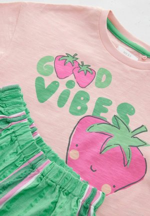 Camiseta rosa con el texto "Good Vibes" y un gráfico de fresa, combinada con pantalones cortos verdes que tienen una cinturilla con volantes y acentos a rayas rosas.