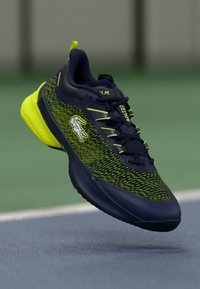 Marineblå og neongul treningssko med teksturert overdel, hvit krokodille-logo og snøring, svevende over en sportsbane.