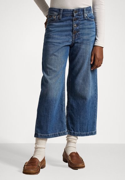Široke traperice u srednje plavoj denimu s izblijedjelim dijelovima, s visokim strukom i pet dugmadi. Uparene s smešnim loafers cipelama i krem čarapama.
