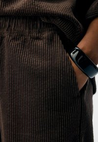 Brun kordfløjlstof med lodrette ribbet tekstur, parret med et skinnende sort armbånd. Synlig lomme-detalje nær taljen.